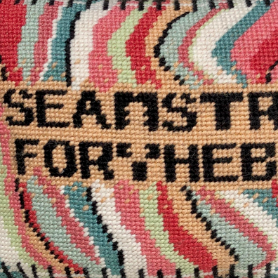 SEAMSTRESS FOR THE BAND stitched in black inside tan box, surrounded by wavy bands of turquoise, pink, red, white, lime and a keyboard border