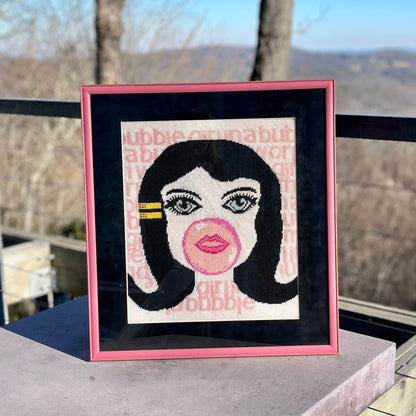needlepoint big-eyed girl blowing pink bubble & hair flipped up, BUBBLE GIRL IN A BUBBLE GUM WORLD text behind image