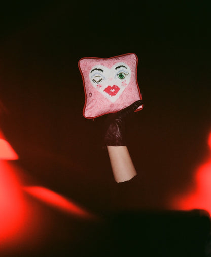 white heart pillow with winking eye and green eye, full lips, pink background with X and O