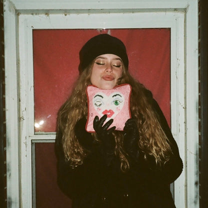 white heart pillow with winking eye and green eye, full lips, pink background with X and O